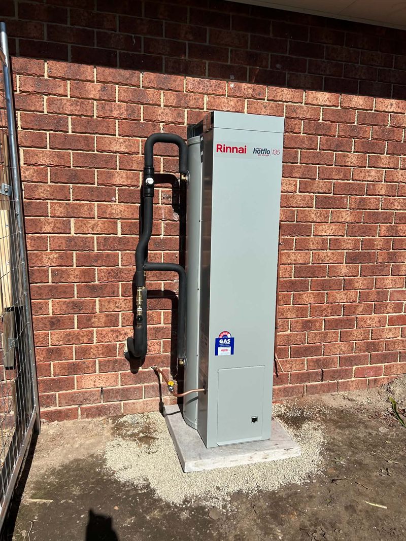 Rinnai Hotflo 135 gas storage hot water unit installed against a brick wall with lagged pipework and a gas compliance tag